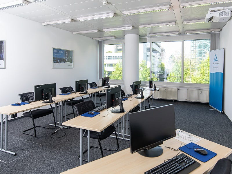 Schulungsraum mit Reihenbestuhlung, Computermonitoren, St&uuml;hlen, zentraler S&auml;ule und Fensterfront mit Tageslicht.