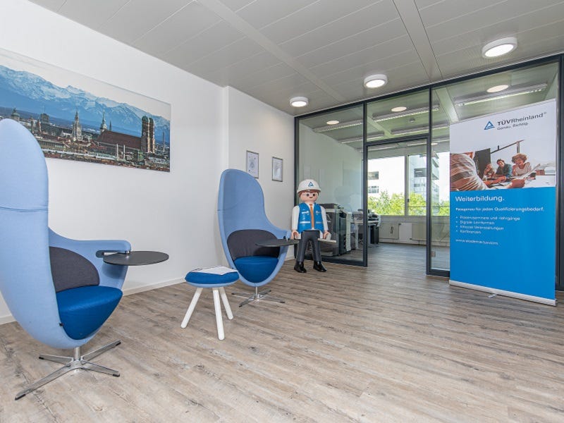 Das Bild zeigt einen modernen Sitzbereich mit zwei blauen Designer-Sesseln und kleinen Beistelltischen. Ein großes Fenster im Hintergrund lässt viel Tageslicht herein. An der Wand hängt ein Bild der Stadtansicht München..