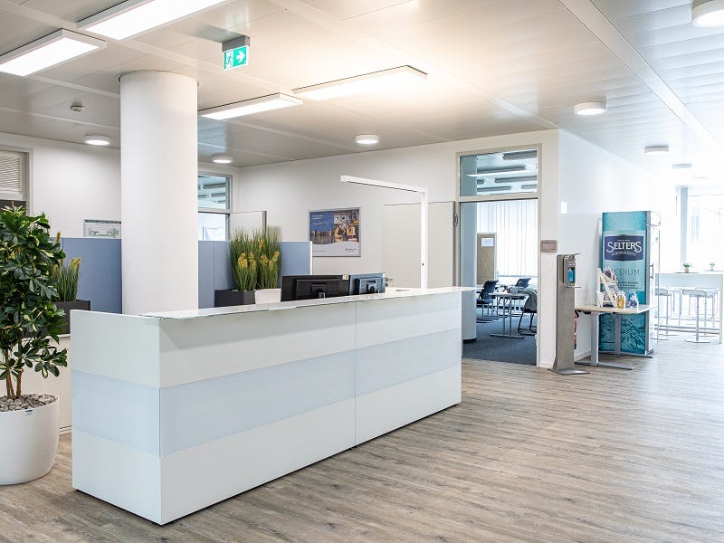 Das Bild zeigt den modernen Empfangsbereich der TÜV Rheinland Akademie München mit einer weißen Rezeption. Pflanzen und moderne Beleuchtung ergänzen das minimalistische und helle Design. Im Hintergrund sind ein Seminarraum und ein Bistrobereich zu sehen.