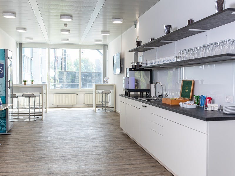 Das Bild zeigt einen Bistrobereich mit weißen Schränken und einer großen Arbeitsfläche. Helle Holzböden und große Fenster, die viel Tageslicht hereinlassen, erzeugen eine freundliche Atmosphäre. Im Hintergrund stehen Tische und Stühle, die als kleiner