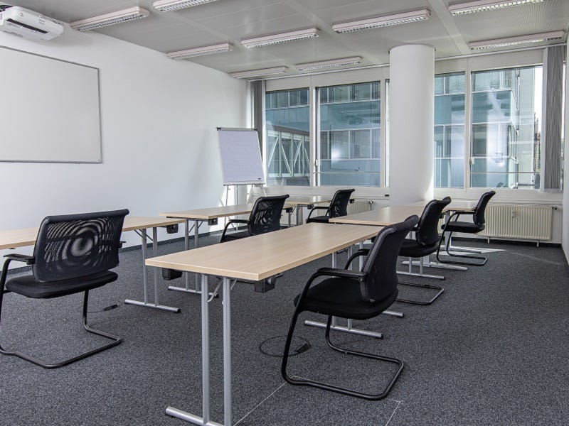 Das Bild zeigt einen modernen Schulungsraum mit weißen Wänden und grauem Teppichboden. Mehrere Tische und schwarze Stühle sind in Reihen angeordnet. Ein Whiteboard und ein Flipchart stehen am vorderen Ende des Raumes, und große Fenster lassen viel Tagesli
