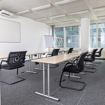 Heller Schulungsraum mit in Reihen angeordneten Tischen, schwarzen St&uuml;hlen, Whiteboard an der Wand und gro&szlig;fl&auml;chigen Fenstern