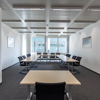Heller Besprechungsraum mit U-f&ouml;rmigem Tischarrangement, schwarzen St&uuml;hlen, S&auml;ule und gro&szlig;fl&auml;chigen Fenstern f&uuml;r viel Tageslicht.