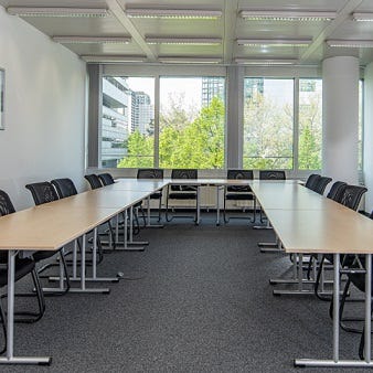 Das Bild zeigt einen modernen Seminarraum mit langen Tischen und schwarzen Stühlen. Große Fenster sorgen für viel natürliches Licht und bieten einen Blick ins Grüne. Die Raumgestaltung ist funktional und schlicht.