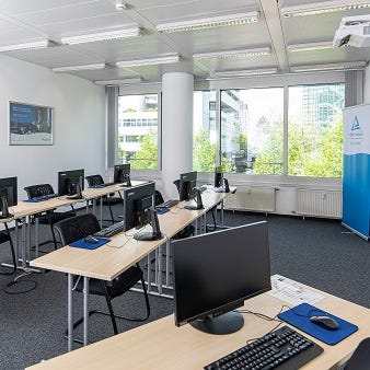 Schulungsraum mit Reihenbestuhlung, Computermonitoren, St&uuml;hlen, zentraler S&auml;ule und Fensterfront mit Tageslicht.