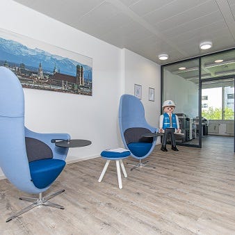 Das Bild zeigt einen modernen Sitzbereich mit zwei blauen Designer-Sesseln und kleinen Beistelltischen. Ein großes Fenster im Hintergrund lässt viel Tageslicht herein. An der Wand hängt ein Bild der Stadtansicht München..