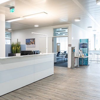 Das Bild zeigt den modernen Empfangsbereich der TÜV Rheinland Akademie München mit einer weißen Rezeption. Pflanzen und moderne Beleuchtung ergänzen das minimalistische und helle Design. Im Hintergrund sind ein Seminarraum und ein Bistrobereich zu sehen.