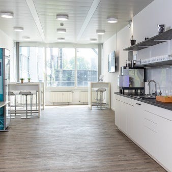 Das Bild zeigt einen Bistrobereich mit weißen Schränken und einer großen Arbeitsfläche. Helle Holzböden und große Fenster, die viel Tageslicht hereinlassen, erzeugen eine freundliche Atmosphäre. Im Hintergrund stehen Tische und Stühle, die als kleiner