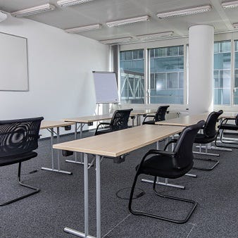 Das Bild zeigt einen modernen Schulungsraum mit weißen Wänden und grauem Teppichboden. Mehrere Tische und schwarze Stühle sind in Reihen angeordnet. Ein Whiteboard und ein Flipchart stehen am vorderen Ende des Raumes, und große Fenster lassen viel Tagesli