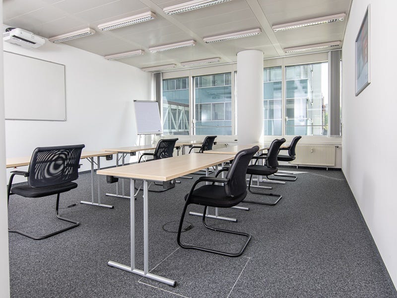 Heller Schulungsraum mit in Reihen angeordneten Tischen, schwarzen St&uuml;hlen, Whiteboard an der Wand und gro&szlig;fl&auml;chigen Fenstern