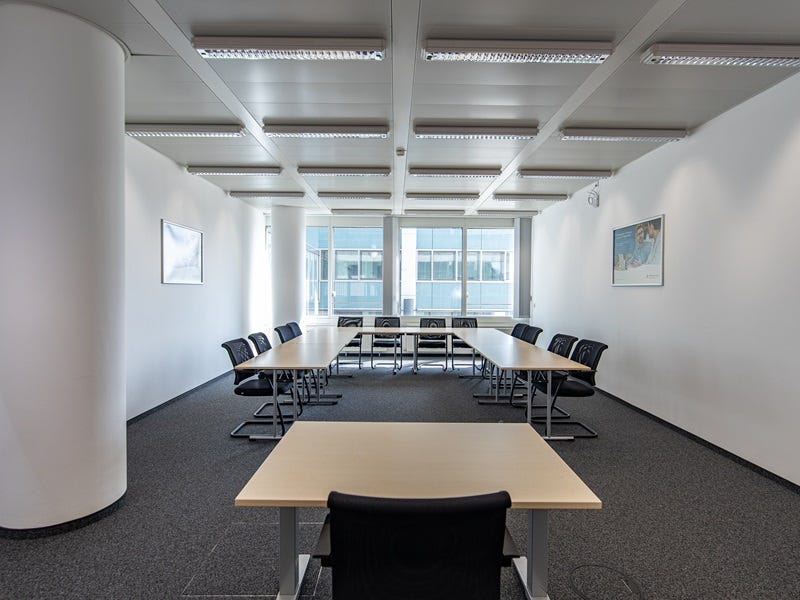 Heller Besprechungsraum mit U-f&ouml;rmigem Tischarrangement, schwarzen St&uuml;hlen, S&auml;ule und gro&szlig;fl&auml;chigen Fenstern f&uuml;r viel Tageslicht.