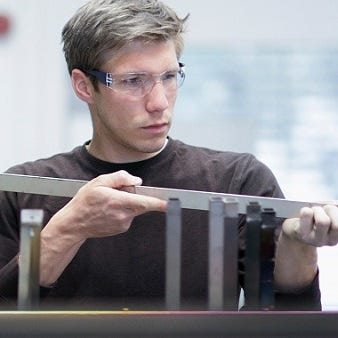 Das Bild zeigt einen Mann, der ein Metallstück betrachtet. Er trägt eine Schutzbrille und prüft, ob das Bauteil präzise gefertigt ist.