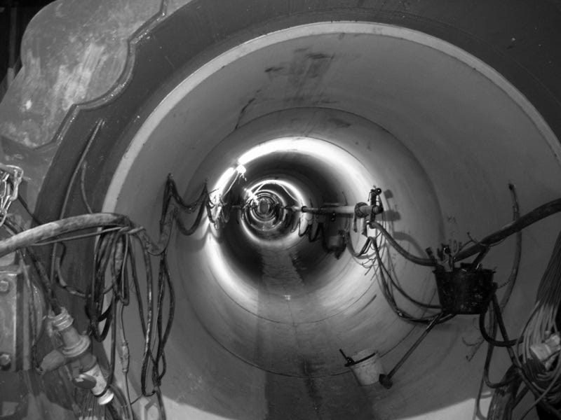 Ein Schwarzweiß-Bild, das das Innere eines großen Rohres oder Tunnels zeigt, mit diversen Kabeln und Rohren, die entlang der inneren Wände verlaufen.