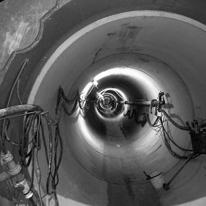 Ein Schwarzweiß-Bild, das das Innere eines großen Rohres oder Tunnels zeigt, mit diversen Kabeln und Rohren, die entlang der inneren Wände verlaufen.