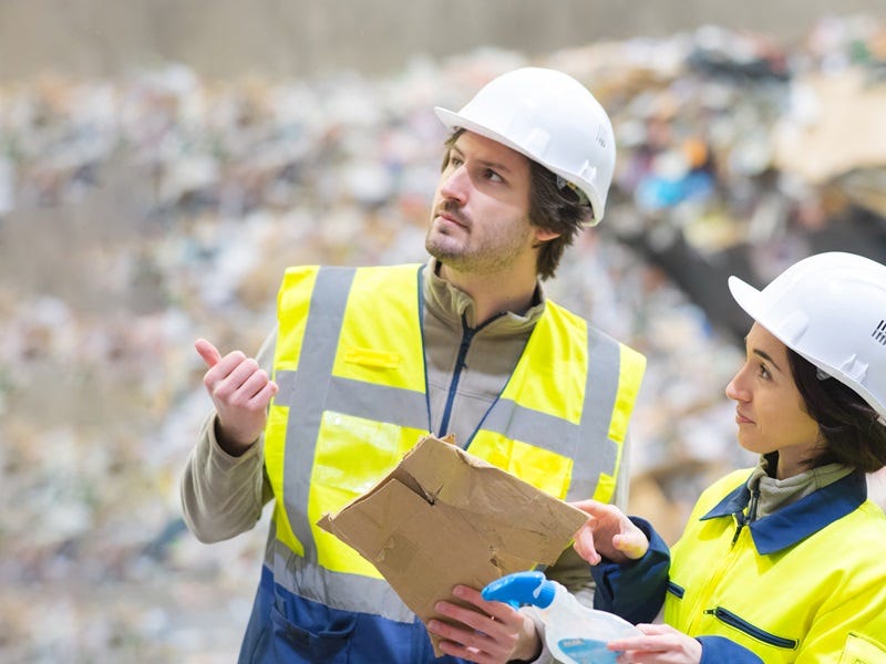 Zwei Fachkräfte mit Warnweste und Schutzhelm inspizieren eine Abfall- oder Recyclinganlage; einer hält Pappe in der Hand, beide schauen prüfend in eine Richtung.