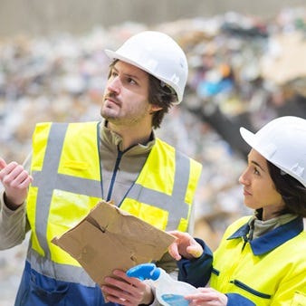 Zwei Fachkräfte mit Warnweste und Schutzhelm inspizieren eine Abfall- oder Recyclinganlage; einer hält Pappe in der Hand, beide schauen prüfend in eine Richtung.