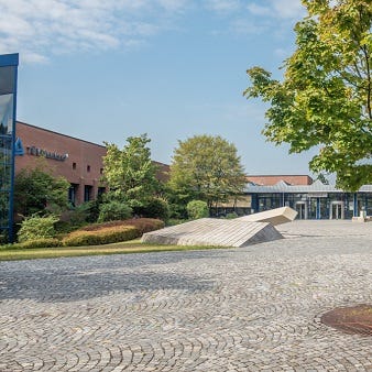 Gepflasterter Vorplatz mit Baumreihe und barrierefreier Rampe vor dem Eingangsbereich des Backsteingeb&auml;udes von T&Uuml;V Rheinland 