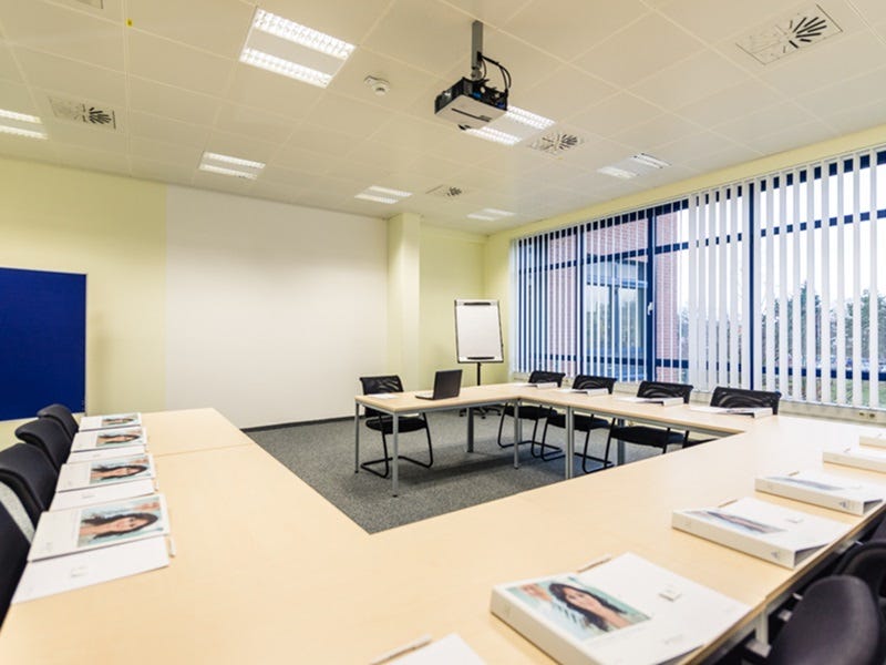 U-f&ouml;rmig angeordneter Tagungsraum mit Tischen und schwarzen St&uuml;hlen, Flipchart, Deckenprojektor und vertikalen Jalousien.