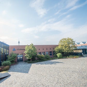 Gebäude der TÜV Rheinland Akademie Nürnberg an einem sonnigen Tag, umgeben von Bäumen und einem gepflasterten Hof. Im Hintergrund der Fernsehturm Nürnberg.