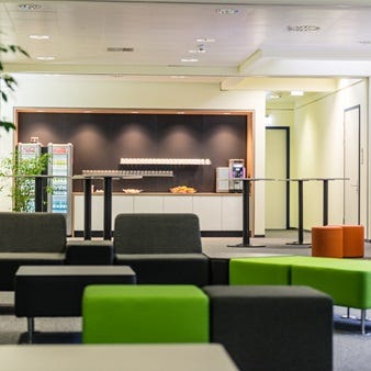 Ein Loungebereich. Im Vordergrund stehen farbenfrohe, bequeme Sitzmöbel in Grün und Grau. Im Hintergrund sind hohe Tische und eine Bar mit Getränken und Snacks zu sehen