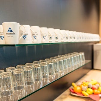 Ein Regal mit mehreren weißen Kaffeebechern, die das Logo der TÜV Rheinland Group tragen, und einer Reihe von Gläsern. Auf der Arbeitsfläche darunter steht eine Schale mit verschiedenen frischen Früchten