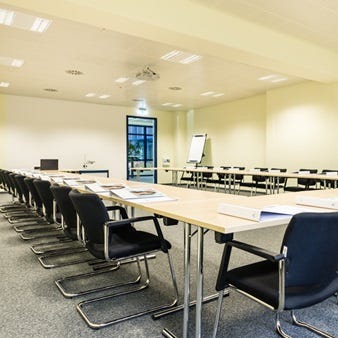 Ein großer Seminarraum mit langen Tischen, geordneten schwarzen Stühlen und einem Präsentationsbildschirm im Hintergrund. Auf den Tischen liegen Blöcke und Stifte für die Teilnehmer bereit