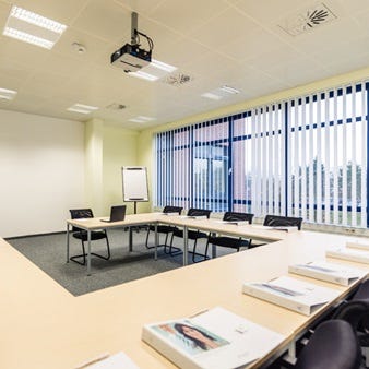 Ein heller und moderner Seminarraum mit großen Fenstern und Jalousien. Der Raum ist mit länglichen Tischen und schwarzen Bürostühlen ausgestattet. Auf den Tischen liegen mehrere Ordner und Broschüren. An der Decke befindet sich ein Beamer.