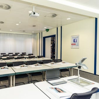 Heller Schulungsraum mit Reihenbestuhlung, Tischen und schwarzen St&uuml;hlen, Deckenprojektor und geschlossenen Jalousien.