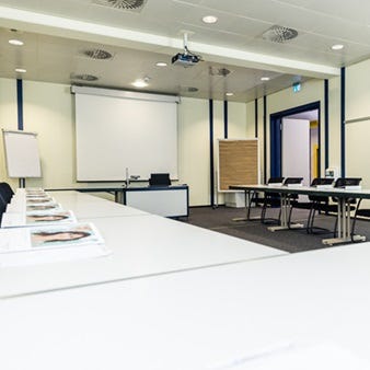 Heller Seminarraum mit in U-Form angeordneten Tischen, schwarzen St&uuml;hlen, Flipchart, Deckenprojektor und Leinwand