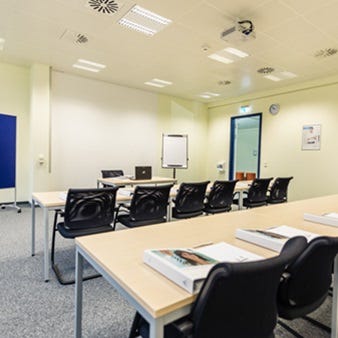 Heller Schulungsraum mit Reihenbestuhlung, schwarzen St&uuml;hlen, Flipchart, Deckenprojektor und Tageslichteinfall durch eine T&uuml;r.