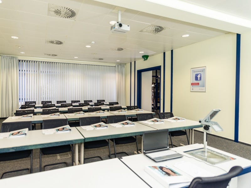 Heller Schulungsraum mit Reihenbestuhlung, Tischen und schwarzen St&uuml;hlen, Deckenprojektor und geschlossenen Jalousien.