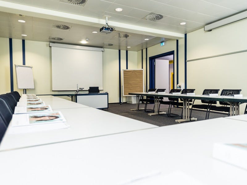 Heller Seminarraum mit in U-Form angeordneten Tischen, schwarzen St&uuml;hlen, Flipchart, Deckenprojektor und Leinwand