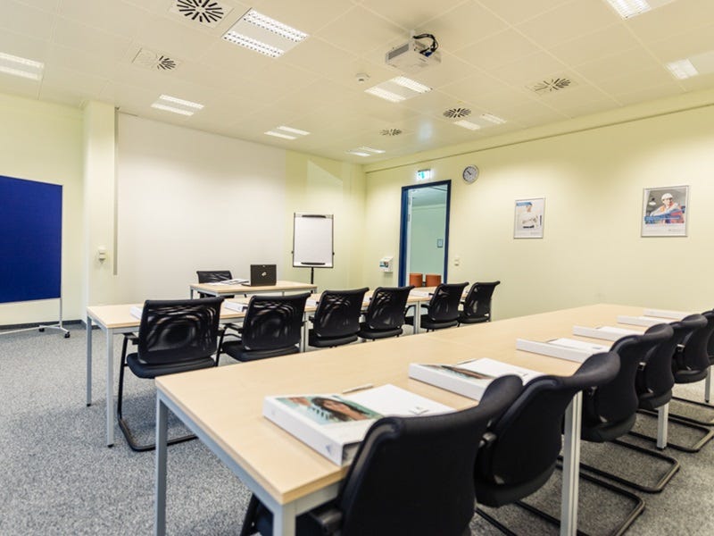 Heller Schulungsraum mit Reihenbestuhlung, schwarzen St&uuml;hlen, Flipchart, Deckenprojektor und Tageslichteinfall durch eine T&uuml;r.