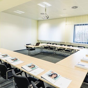 U-f&ouml;rmiger Tagungsraum mit hellem Tischarrangement, schwarzen St&uuml;hlen, Flipchart, Deckenprojektor und vertikalen Jalousien.
