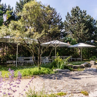 Terrasse mit wei&szlig;en Gartenm&ouml;beln und Sonnenschirmen, umgeben von B&auml;umen und &uuml;ppiger Bepflanzung