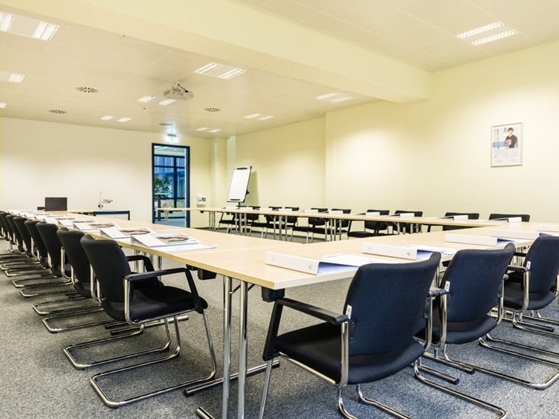 Heller Seminarraum mit langen Tischen und schwarzen St&uuml;hlen, Flipchart und Deckenprojektor.