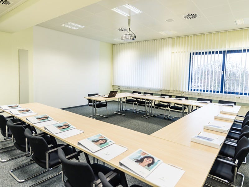 U-f&ouml;rmiger Tagungsraum mit hellem Tischarrangement, schwarzen St&uuml;hlen, Flipchart, Deckenprojektor und vertikalen Jalousien.