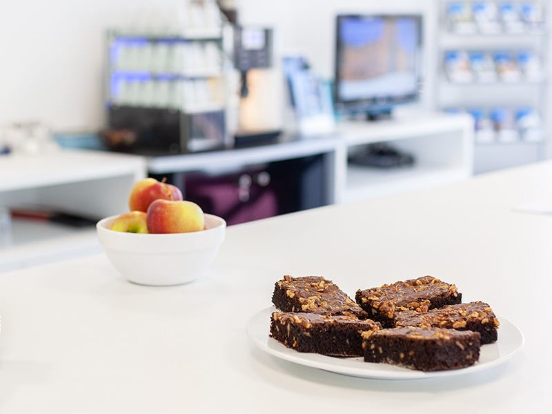 Tisch mit Teller voll Brownies und Schale mit &Auml;pfeln vor Kaffeemaschine und TV-Bildschirm in hellem Loungebereich