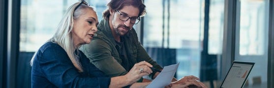 Frau mittleren Alters mit Blatt Papier in der Hand sitzt mit dunkerlhaarigem Mann mit Brille an einem Schreibtisch vor einem Laptop und bespricht etwas