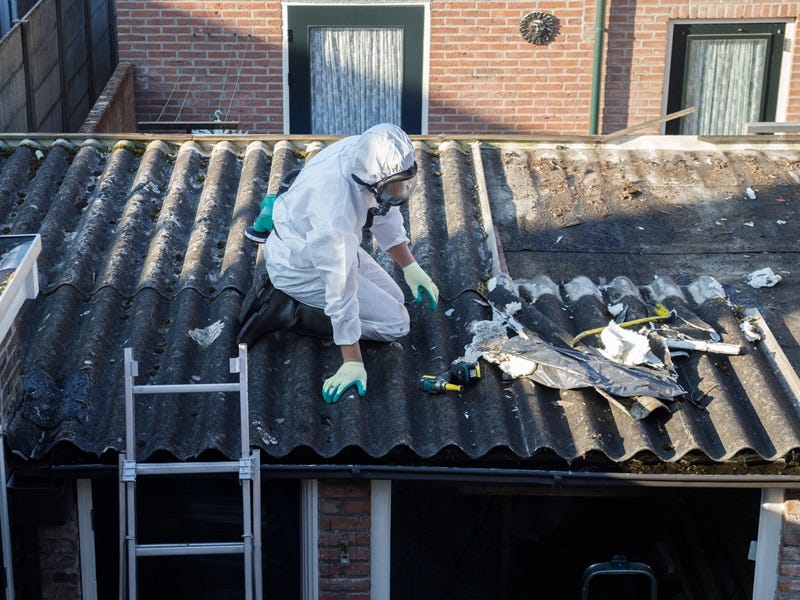 Arbeiter in Schutzkleidung entfernt asbesthaltige Dachplatten auf einem Wellplattendach eines Wohngebäudes.