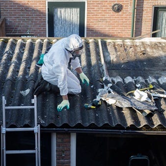 Arbeiter in Schutzkleidung entfernt asbesthaltige Dachplatten auf einem Wellplattendach eines Wohngebäudes.