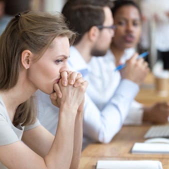 Verärgerte, frustrierte und verwirrte Mitarbeiterin, die ihre Hände unter dem Kinn verschränkt, sitzt in einem modernen Großraumbüro mit multinationalen Kollegen und wirkt ratlos.