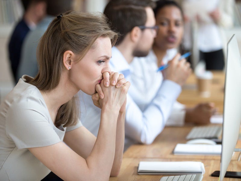 Verärgerte, frustrierte und verwirrte Mitarbeiterin, die ihre Hände unter dem Kinn verschränkt, sitzt in einem modernen Großraumbüro mit multinationalen Kollegen und wirkt ratlos.