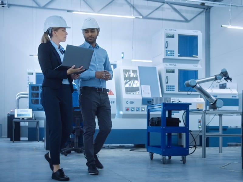 Das Bild zeigt zwei Personen in einem modernen Industriebetrieb. Beide tragen Schutzhelme und formelle Arbeitskleidung. Die Frau hält einen Laptop, während der Mann mit ihr spricht und ebenfalls auf den Bildschirm blickt. 
