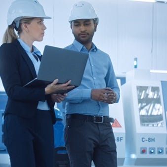 Das Bild zeigt zwei Personen in einem modernen Industriebetrieb. Beide tragen Schutzhelme und formelle Arbeitskleidung. Die Frau hält einen Laptop, während der Mann mit ihr spricht und ebenfalls auf den Bildschirm blickt. 