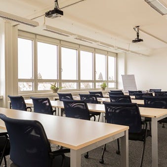 Seminarraum mit mehreren Reihen aus hellen Tischen und blauen St&uuml;hlen, Deckenprojektor, Flipchart und gro&szlig;en Fenstern.