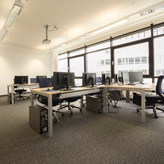 Seminarraum mit mehreren PC-Arbeitspl&auml;tzen, Desktop-PCs und Monitoren, Deckenprojektor, gro&szlig;en Fenstern und gr&uuml;ner Garderobe.