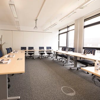 U-f&ouml;rmige Bestuhlung in einem hellen Schulungsraum mit blauen St&uuml;hlen, Deckenprojektor und gro&szlig;en Fenstern.