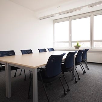 Heller Besprechungsraum mit rechteckigem Tisch, acht blauen St&uuml;hlen, gro&szlig;em Fenster, Pflanze und Garderobenst&auml;nder.