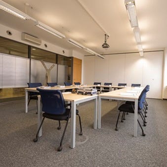 Heller Schulungsraum mit U-f&ouml;rmigen Tischen, blauen St&uuml;hlen, Deckenprojektor und gro&szlig;en Fenstern f&uuml;r Seminare und Meetings.